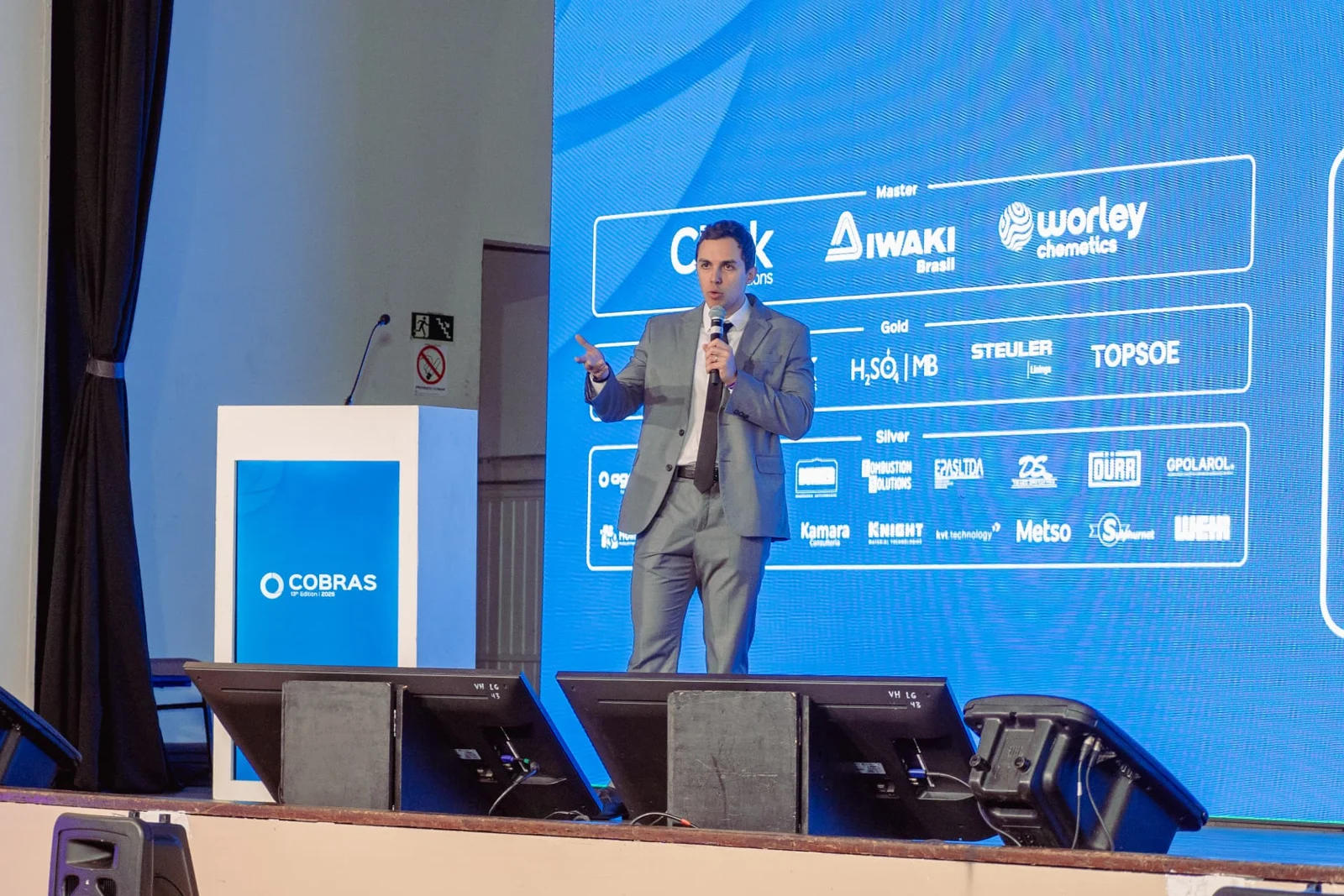 Palestrante no palco diante de painel com marcas patrocinadoras